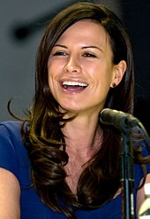 A seated woman with brown hair extending below her shoulders and wearing a blue shirt smiles as she looks to her right.