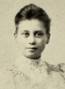 A young white woman with dark hair brushed back from her forehead, wearing a high-collared white lacy blouse or dress