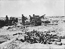 Uma arma de artilharia e sua tripulação em um deserto. Uma pilha de caixas está em primeiro plano.