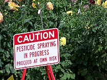 This is in Manito Park in Spokane, WA. It is a red-and-white caution sign warning of pesticide usage. "Caution: Pesticide Spraying. Enter at your own risk" is written.