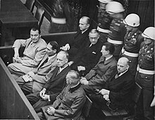 Photo noir et blanc prise en 1946, au procès de Nuremberg. Dans le box des accusés sont assis huit hommes, sur deux rangées de quatre. Au premier rang (de haut en bas) : Hermann Göring, Rudolf Heß, Joachim von Ribbentrop et Wilhelm Keitel en uniforme. Au deuxième rang (de haut en bas) : Karl Dönitz, Erich Raeder, Baldur von Schirach et Fritz Sauckel. Derrière eux (en haut, à droite), se tiennent quatre membres de la police militaire, debout, les mains derrière leur dos.