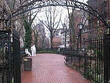 A color photograph of Christopher Park in winter, showing the wrought iron entrance arch in the foreground and the brick pavement surrounded by five and six-story brick buildings; in the center background are four white statue figures: two males standing, one with his hand on the other's shoulder and two females seated on a park bench, one woman with her hand touching the other's thigh. All are dressed in jeans and loose clothing