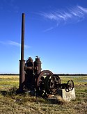 Photo of a very rusted industrial-looking pump with tall tube and wheel sits in a flat rice field.