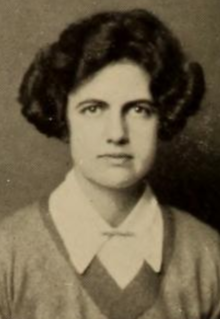 A young white woman with short dark thick hair, wearing a v-necked sweater over a collared shirt, pinned at throat