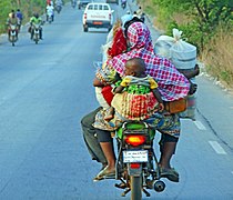 Une femme avec un bébé, des bagages dans les deux bras sur moto sur la route de Parakou vers son village