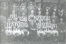 A football team comprising ten players in striped shirts and one in a shirt of a single colour pose for the camera. Five of the men are standing and the other six seated in front of them. Also posing with them are an elderly man in a bowler hat with a chain of office around his neck, and twelve men in business suits, some of whom are wearing hats. A crowd of spectators is visible behind the group.