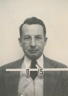 Mug shot of a man with glasses in suit and tie. The number O-15 appears in front of him.