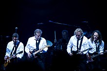 A color photograph of four members of the Eagles on stage with guitars