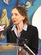 Color photo of an elderly women at a podium, speaking into a mic.