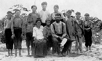 Outdoor scene of a man and woman seated on chairs in front of a group of ten children of varying ages, barefoot and wearing simple clothing