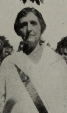 An older white woman, wearing a white dress with a ribbon sash