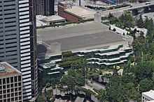 An aerial view of a concrete building with a bare roof and several terraced glass walls, abutting a park full of trees between several high-rise towers