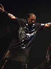 Чоловік тримає мікрофон у правій руці, перебуваючи на сцені. Він одягнений у чорну футболку з тигровим обличчям на ній та вузькі шкіряні штани.