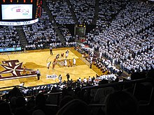 a crowd of people in mostly white T-shirts watch an indoor basketball game; a T with a start on it is upside down and featured prominently under the players