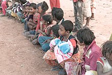 Row of young children sitting or squatting in the dirt wearing dirty clothing, smeared in excrement, one child clutches two towels
