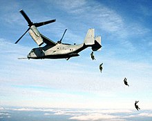 Four U.S. Marine paratroopers jump from the rear loading ramp of an MV-22 Osprey.
