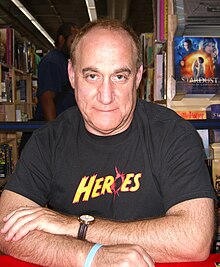 A middle-aged white man is sitting in a bookstore, wearing a "Heroes"-branded t-shirt; his arms are crossed, he is facing and looking into the camera with a neutral expression.