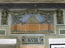 A faience mosaic on the wall that depicts a New Amsterdam step-gabled house with the palisade wall in front of it. The mosaic is topped by blue faience swags. There is a faience cornice with scrolled and foliate detail.