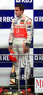 Fernando Alonso dons white fireproof overalls as he stands on a podium with a bottle of champagne