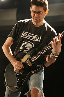 A frowning man with glasses playing a black guitar, wearing a black t-shirt and ripped blue jeans.