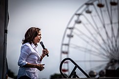 Kobieta z ciemnobrązowymi włosami do ramion ubrana w białą koszulę i trzymająca mikrofon na tle diabelskiego młyna.