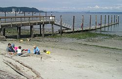 The public pier and waterfront at Manchester during a summer low tide. Bainbridge Island is in the background on the left, and Seattle is far away on the right.