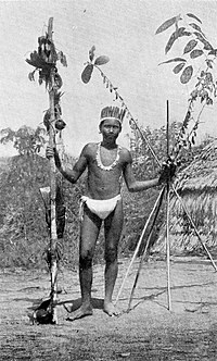 Black and white photograph of a man in a loincloth and feathers with a pole in one hand and four long sticks in the other.