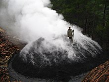 An aerial view of a bearded man in a coat, with a handmade rake, standing on a low, ~10m wide, rounded mound of black earth. The mound is emitting many slender streamers of smoke from all of its surface except the bottommost meter. The steamers merge and stream diagonally away from the viewer on a gentle but chill morning breeze, making an opaques white cloud. The visible background is dark coniferous forest; in the foreground, a glimps of another pile, this one tightly packed wood not yet covered with soil.