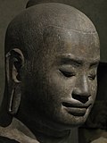 ジャヤーヴァルマン7世頭部像　バイヨン期様式　12世紀　アンコール遺跡群、タ・プローム出土？　ギメ東洋美術館蔵　アンコール時代には、国王自身が信奉する宗教の最高神（仏教においては仏教尊格）の化身であり、死後は帰一するというデーヴァ・ラージャ（神王信仰）が行われた[304]。