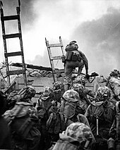 photograph of Marines waiting to climb the seawall at Inchon, while one Marine centered is bent over while dismounting the top of a ladder