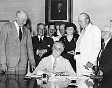 Franklin D. Roosevelt sits at a desk signing a document. Several people stand watching behind him.
