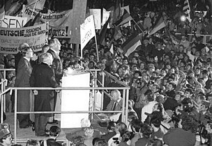 Černobílá fotografie zachycující zábradlím ohrazené pódium s několika řečníky obklopené davem s vlajkami a transparenty