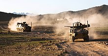 Colour photo of five military armoured fighting vehicles driving through dusty terrain