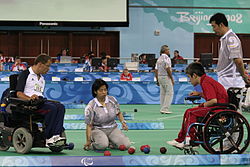 Dois atletas em cadeira de rodas estão frente-a-frente, ambos olhando para o chão, onde estão as bolas usadas no esporte. Uma juíza está agachada entre eles e outro observa em pé, ao fundo.