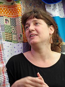 A white woman with brown hair in a black shirt looks upwards and to her right, standing in front of a colorful quilt.