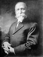 Photographie noir et blanc en buste de trois-quart d'un homme blanc d'âge mûr, front dégarni, aux larges moustaches et bouc, bras croisé sur sa veste