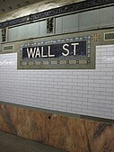 View of the wall along the platform. There is a a pink marble wainscoting on the lowest part of the wall, with white glass tiles above. A white-on-blue tile plaque with the words "Wall Street" and floral motifs is also placed on the wall.