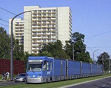 Barevná fotografie s pohledem na modrou tramvaj projíždějící pod výškovým panelovým domem