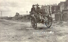 Two men in sombreros riding in a donkey-cart with a line of feet sticking out the back. They are riding down a dirt street away from the camera, with a line of buildings on the right. Dated 15 May 1911.