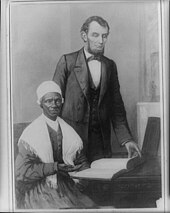 Photographie noir et blanc d'une femme noire assise qui montre de la main une bible et un homme blanc, le président des États-Unis, debout derrière elle
