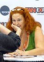 A woman with red hair, glasses atop her head, and a green shit sitting with one of her hands on her mouth.