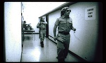 Black and white photo of a white hallway with a plaque reading "Stanford County Prison". Two white male guards in uniforms wearing sunglasses walk along the hallway. One white male guard is sitting on top of a table in a corner in the background.