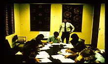 Photo of a seminar room with a large table. Eight white male people are sitting at the table filling out papers spread out on the table. Another white male is standing at the side observing them.