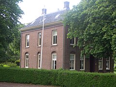 A red brick house with several windows is behind green trees.