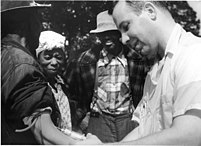 A white doctor has inserted a large needle to draw blood from a black man who stands with his arm at his side. The photo is in black and white. The black man is on the right while the white man stands to the left of him in the frame. Two other black Americans gaze down at the black man's arm as his blood is drawn.