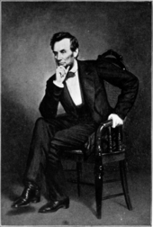 Photographie noir et blanc en pied et assis vers l'avant d'un homme blanc aux cheveux noirs, une main soutenant son menton, l'autre appuyée sur un bras de fauteuil, jambes croisées