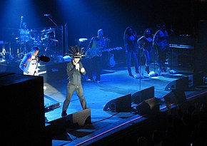 A band performing on stage; a male singer wearing a head-dress, along with a guitarist, a drummer, a bassist and three female vocalists