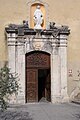 この教会の左側の扉には、上部のパネルに小さなカルトゥーシュが描かれ、そこから落下する典礼用のトロフィーがぶら下がっている。