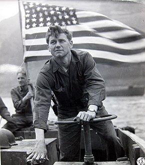 A sailor mans the helm of an LCVP. Old Glory flies in the background.