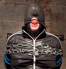A man is chained up and hooded so he can't see. A large red ball is strapped firmly in his mouth so he can't speak.